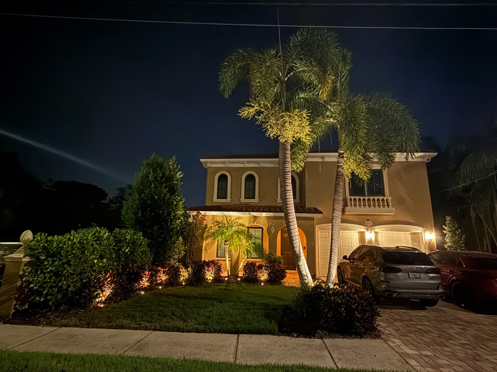 Two-story Tampa home at night with uplit palms, lit garden beds, and path lights along the walkway, a real LogiBuilt install