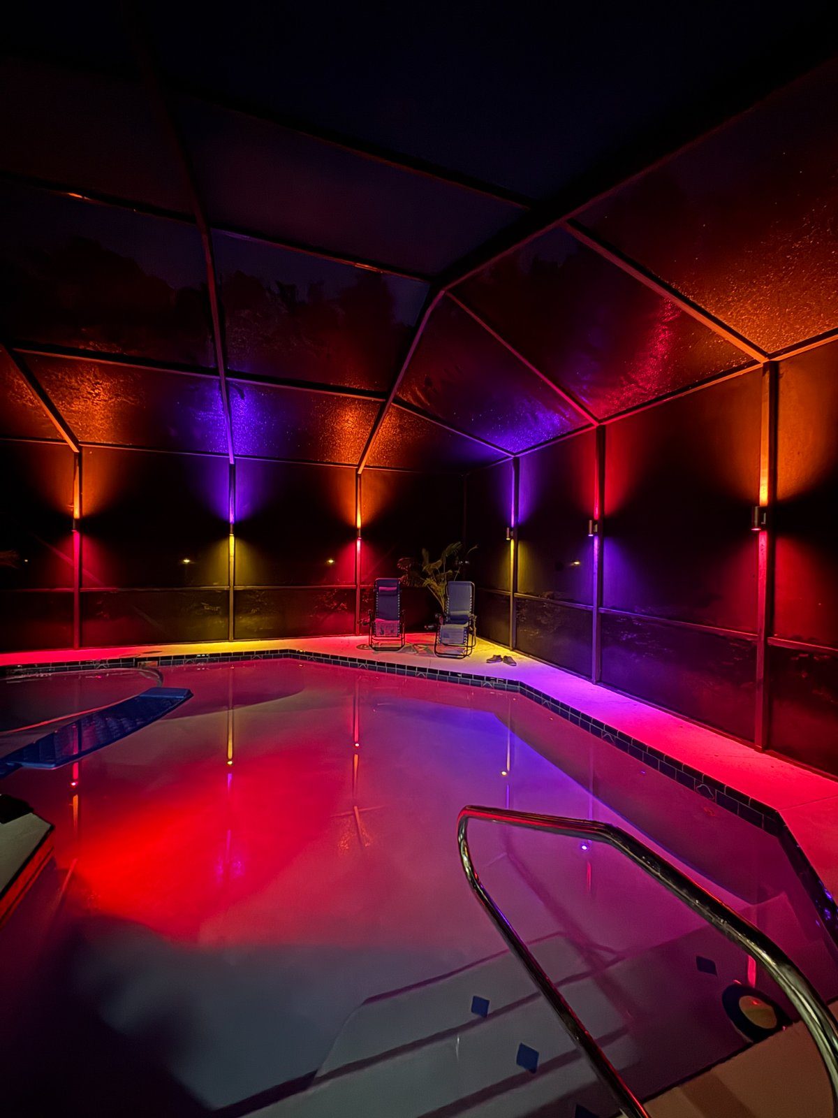 Pool cage at night with color sconces alternating red and blue on every post, reflected in the pool