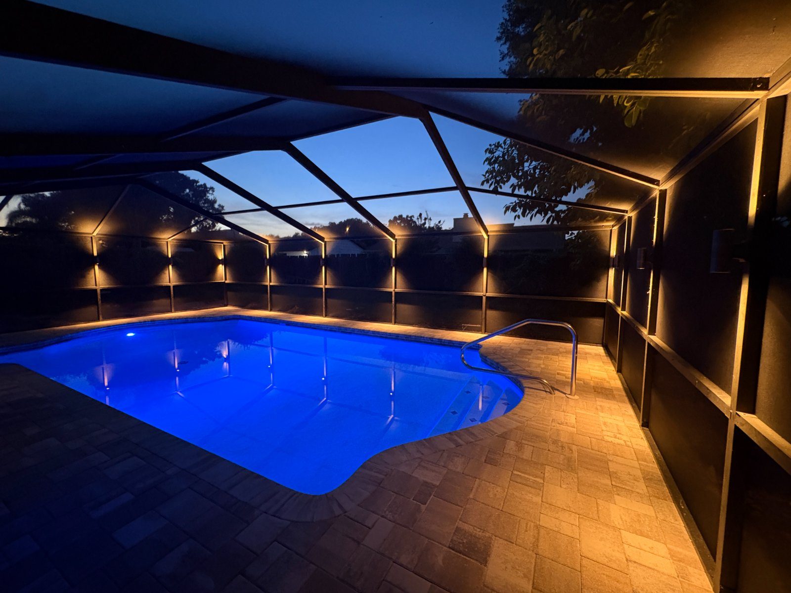 Tampa Bay pool cage at dusk with warm brass up and down sconces lit on every screen post and the pool glowing blue