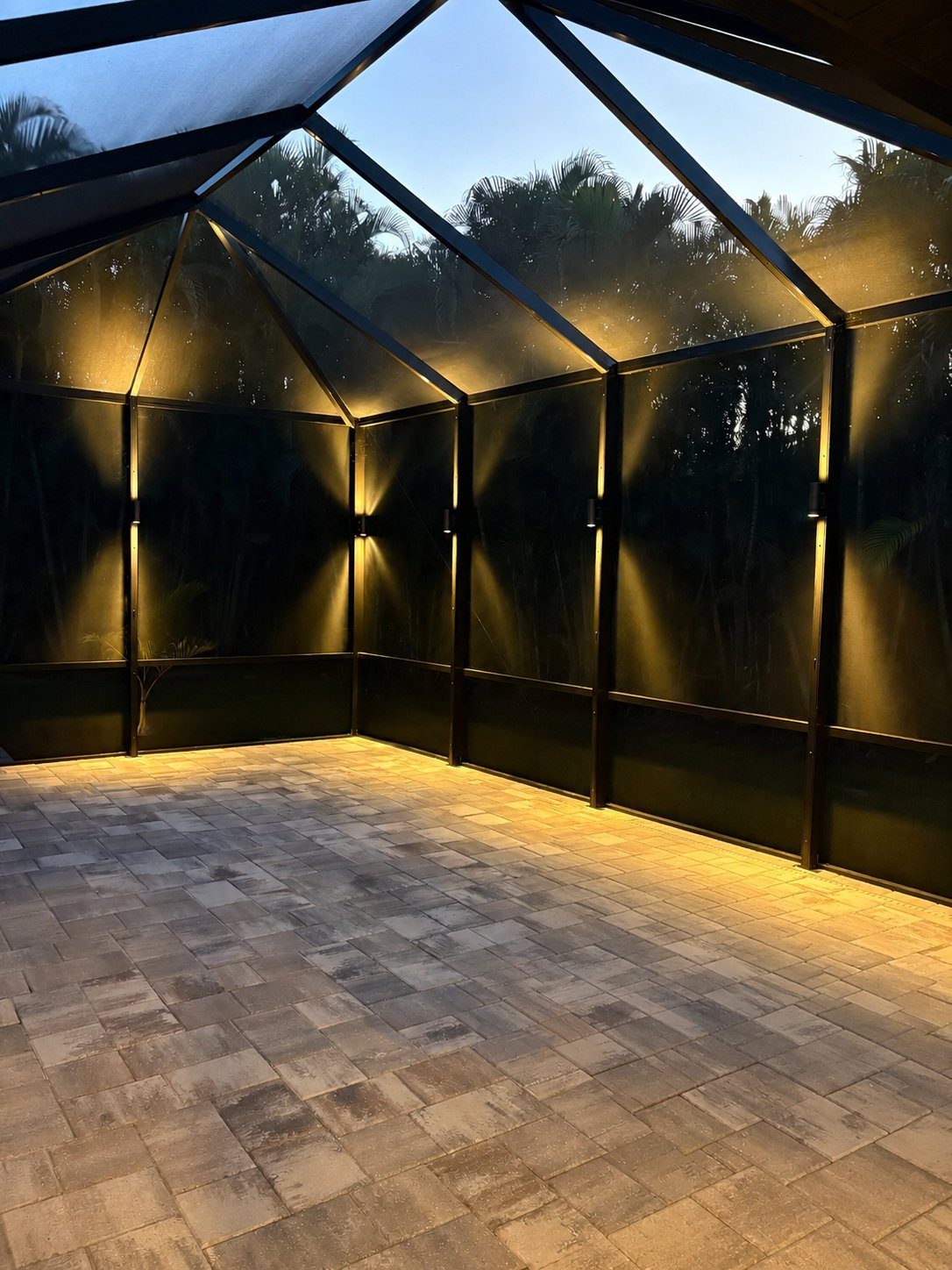 Inside a screened lanai at dusk, brass sconces on each post casting up and down cones of warm light onto the paver deck