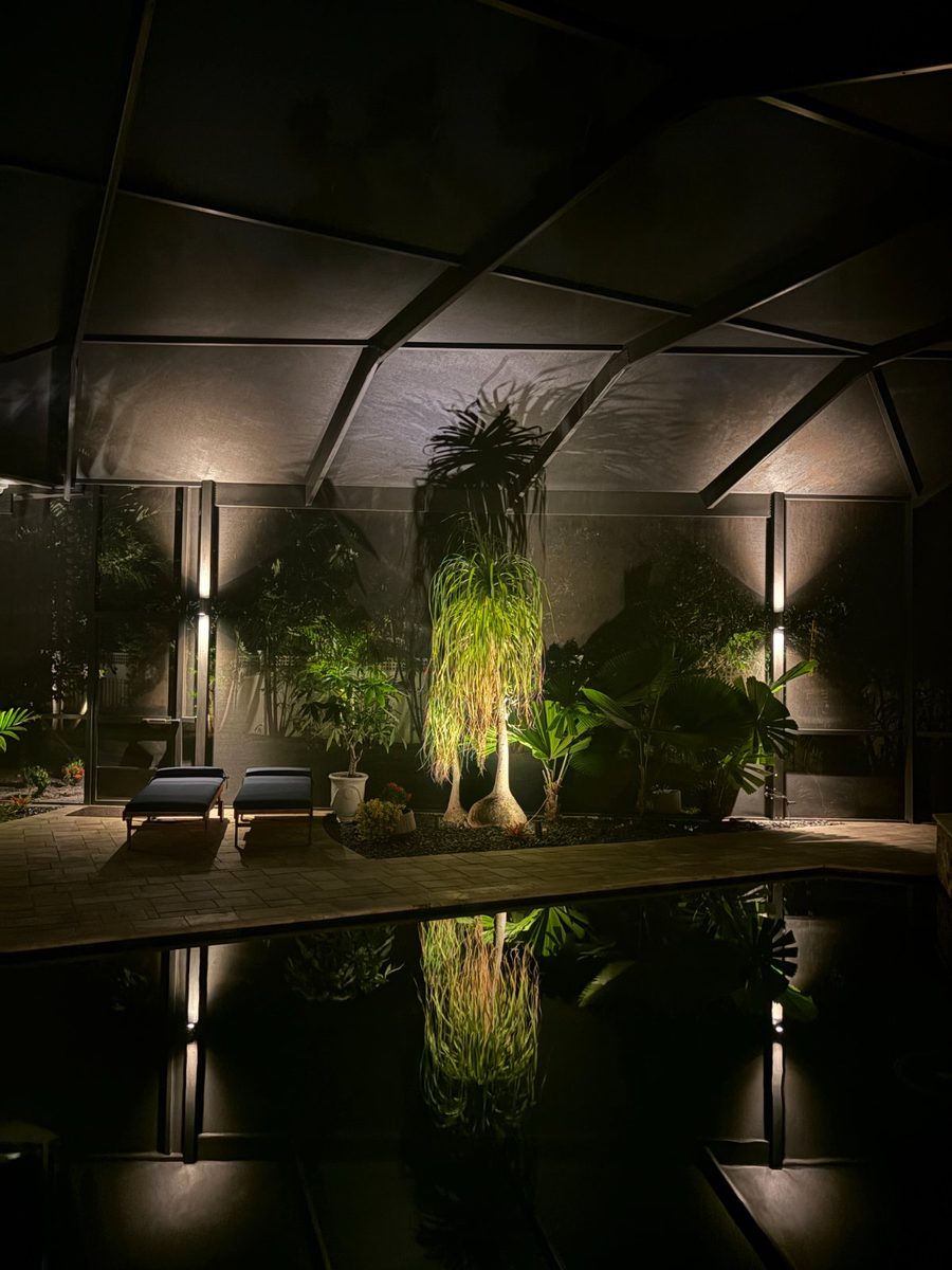 Screened lanai at night with sconces on the posts, a planted bed uplit, and reflections in the pool